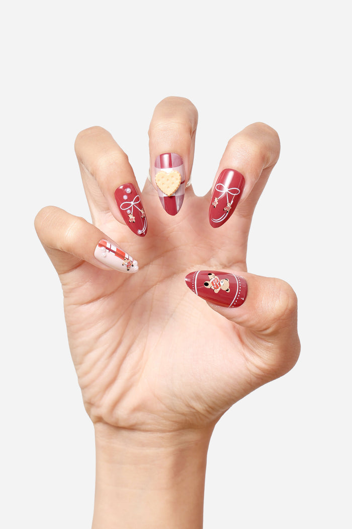 Red almond press-on nails with bows, bear charms, pearls, and plaid accent nail with cookie.
