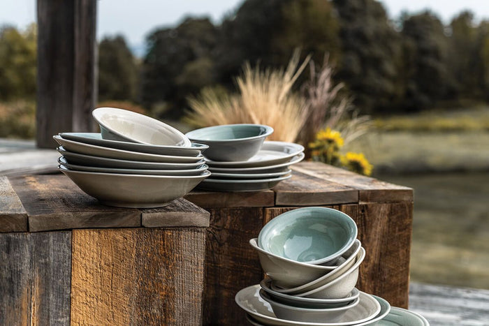 Stacked celadon and cream dinnerware bowls displayed on weathered wood surfaces outdoors.