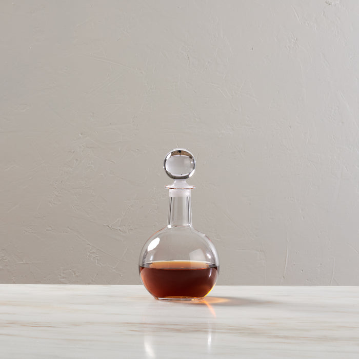 Clear glass decanter with spherical body, narrow neck, and spherical stopper, filled partially with amber liquid.
