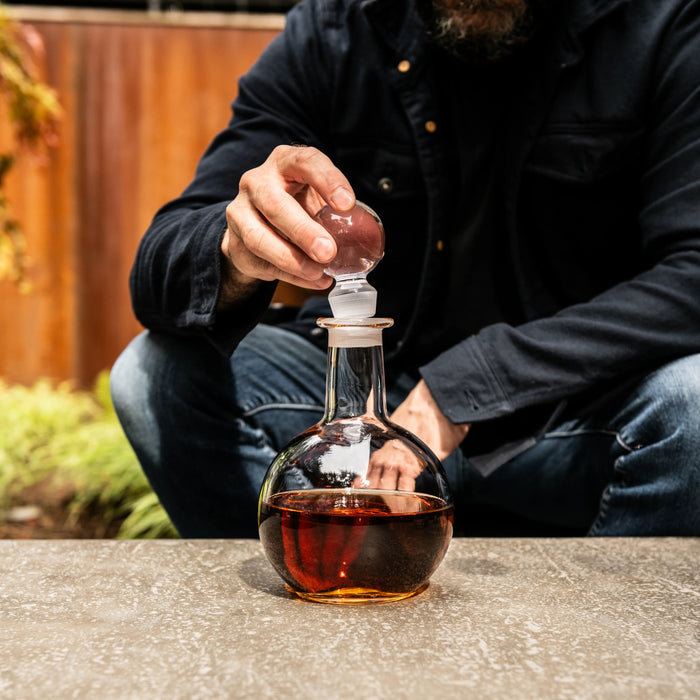 Clear glass decanter with stopper filled with amber liquid, held by a person in dark jacket.