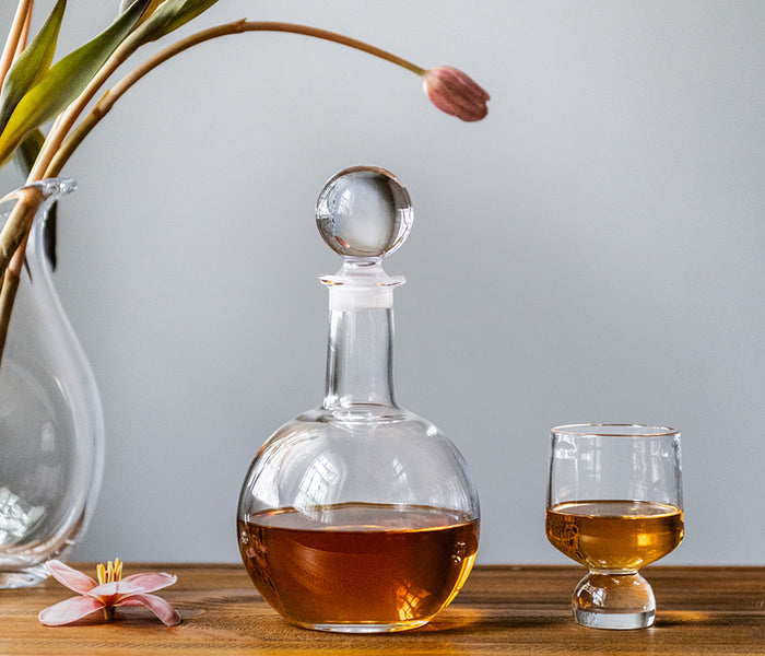 Clear glass decanter with spherical stopper filled with amber liquid, displayed with matching glass and decorative flowers.