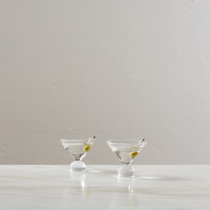 Two martini glasses filled with clear liquid and green olives on white pedestals against a textured beige wall.