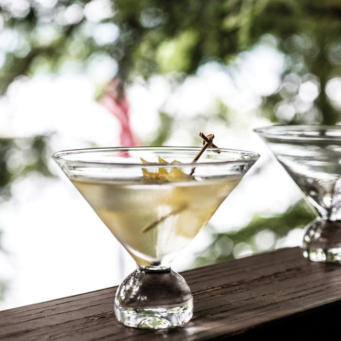 Two martini glasses filled with pale liquid and olive garnish on wooden surface outdoors.