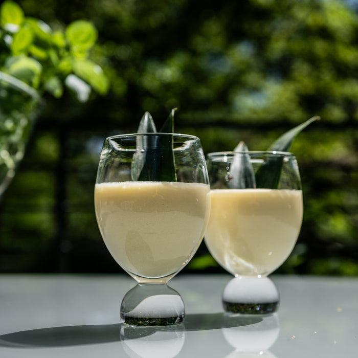 Two clear glass tumblers with white bases filled with pale beverage and black stirrers, displayed outdoors.