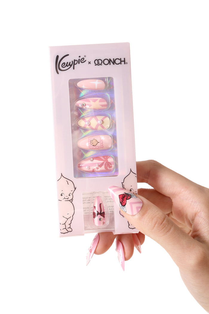 Pink press-on nails with Kewpie and bow designs, packaged and held by a hand.