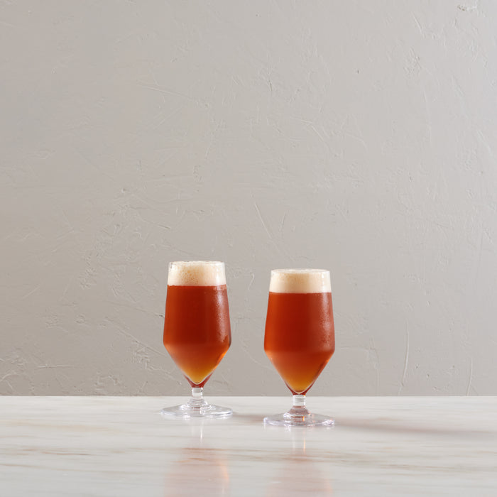 Two clear glass beer glasses filled with amber liquid and foam heads on a white surface.