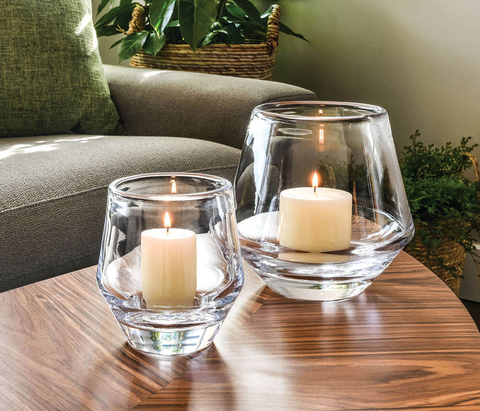 Two clear glass hurricane candle holders with lit cream pillar candles on a wood table.