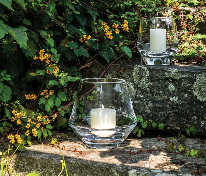 Two clear glass hurricane candle holders with white pillar candles positioned on a moss-covered stone wall surrounded by yell