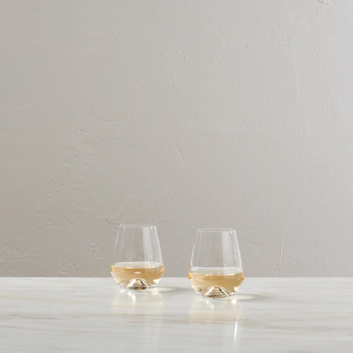 Two clear stemless wine glasses filled with white wine on a white surface.
