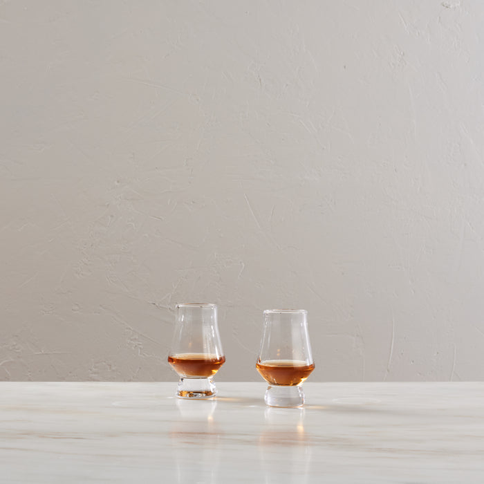 Two clear tasting glasses with tulip-shaped bowls containing amber liquid, displayed on a white surface.