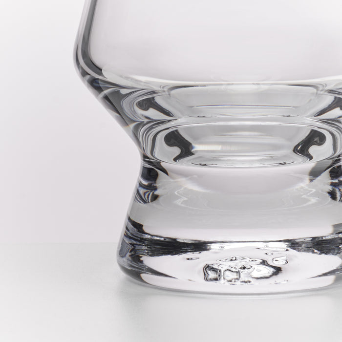 Close-up of a clear glass tumbler with a thick, rounded base and subtle ridged detailing, photographed on a clean white background.