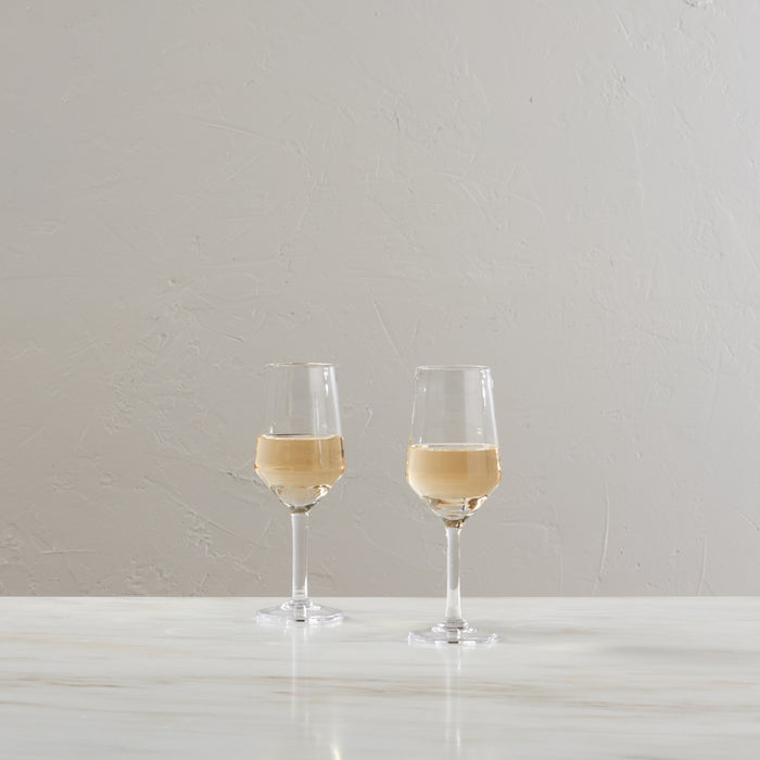 Two wine glasses filled with pale yellow wine on a white surface against a textured light gray wall.