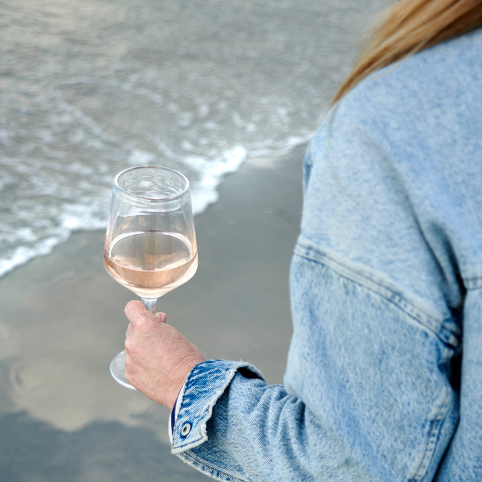 Person holding glass of pale wine on a beach with ocean waves in background.