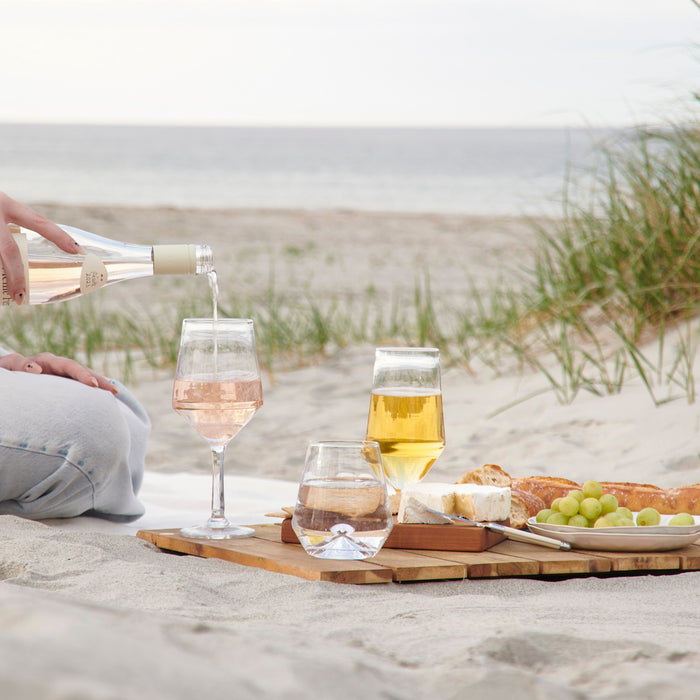 Two clear stemless wine glasses on a beach, one being filled with rosé wine from a bottle.
