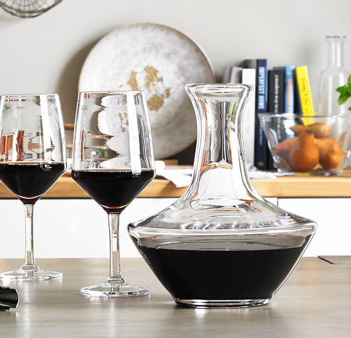 Clear glass decanter filled with red wine, displayed with two matching wine glasses on wooden surface.