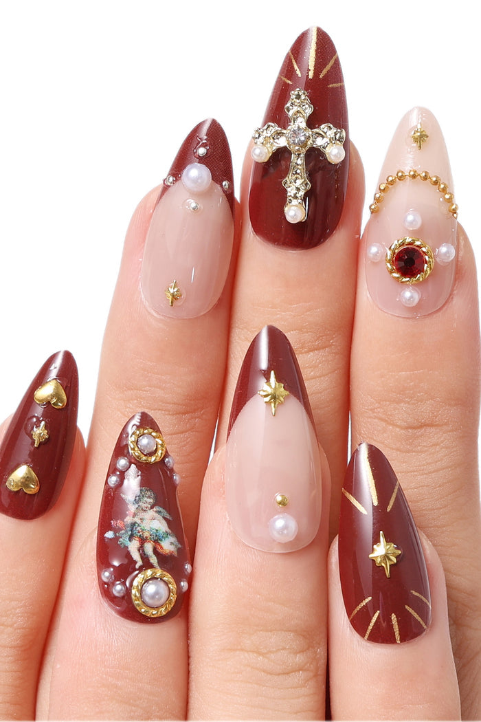 Burgundy French tip press-on nails with gold accents, cherub decals, pearls, and a crystal cross.