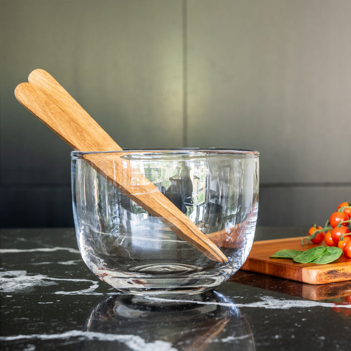 Clear glass bowl with wooden pestle inside, placed on dark countertop next to cutting board with tomatoes and basil.