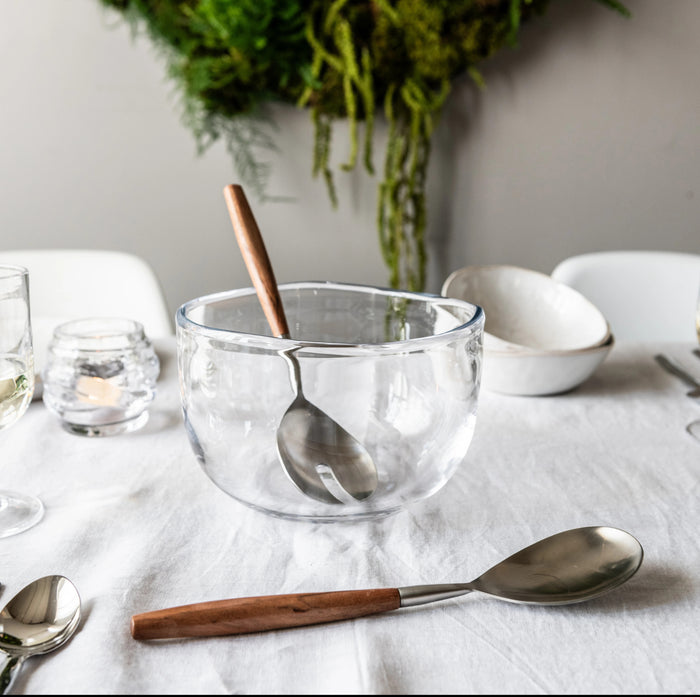 Clear glass bowl with wooden and metal spoon inside, displayed on white table with place settings.