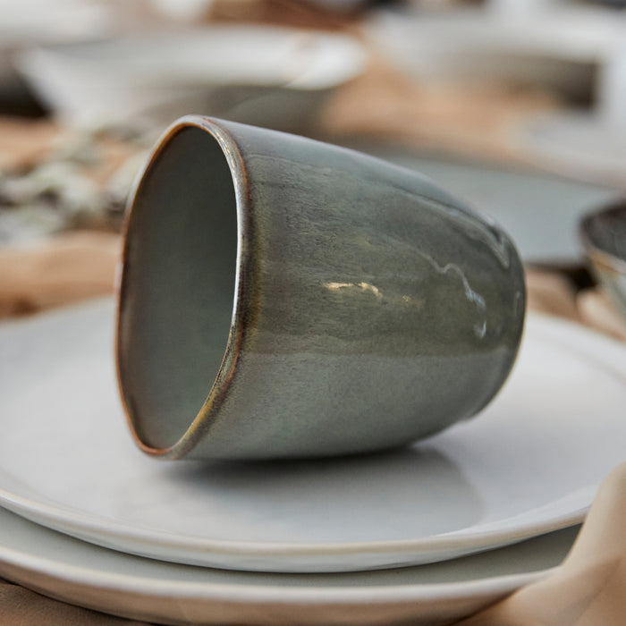 Moss green ceramic cup with rustic rim sitting on stacked white and neutral-toned plates.