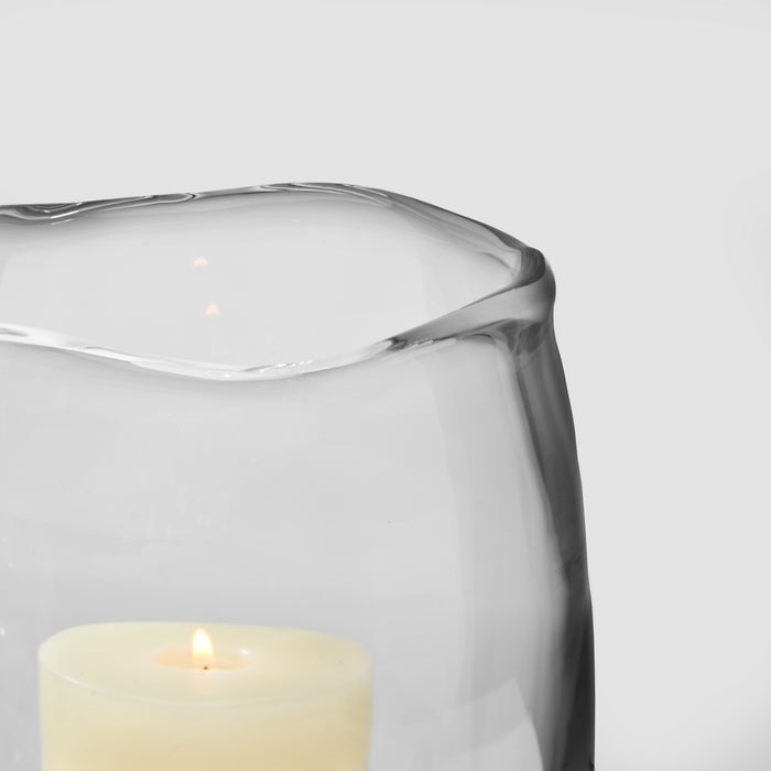Clear glass hurricane with cream-colored candle inside against white background.