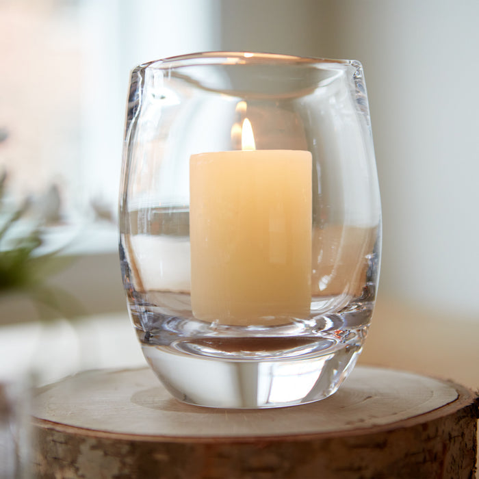 Clear glass hurricane votive holder with cream pillar candle inside, displayed on wooden base.