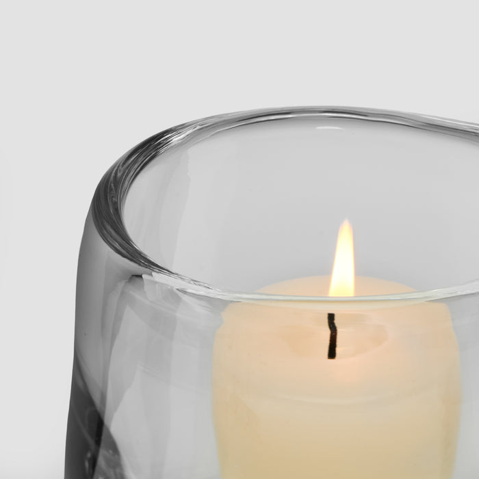 Cream-colored lit candle in clear glass hurricane holder with black wick and protective wrapping.