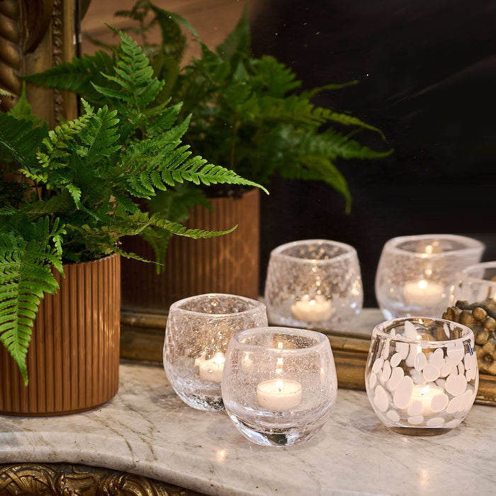 Five clear glass tealight holders with lit candles, displayed on marble surface next to fern plant in bronze vase.