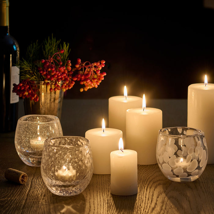 Cream pillar candles with lit flames surrounded by textured glass tealight holders on wooden surface.