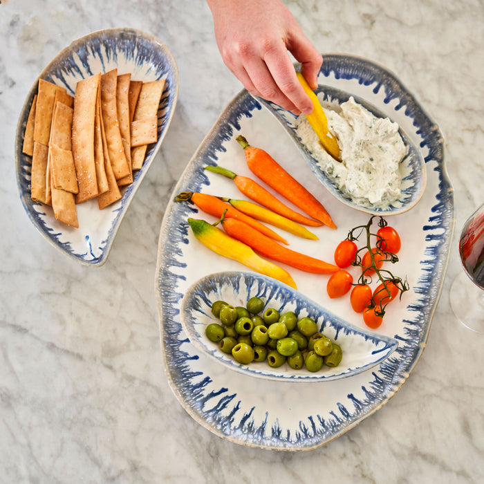 Blue and white striped mussel shell-shaped serving bowls holding crackers, vegetables, olives, and dip on marble surface.