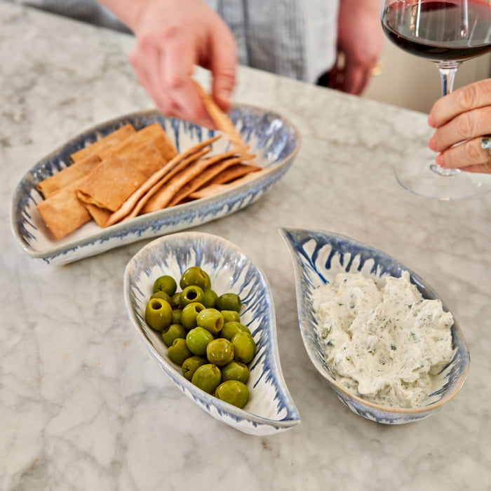 Three shell-shaped bowls in blue and white with olives, cheese dip, and crackers on a marble table.