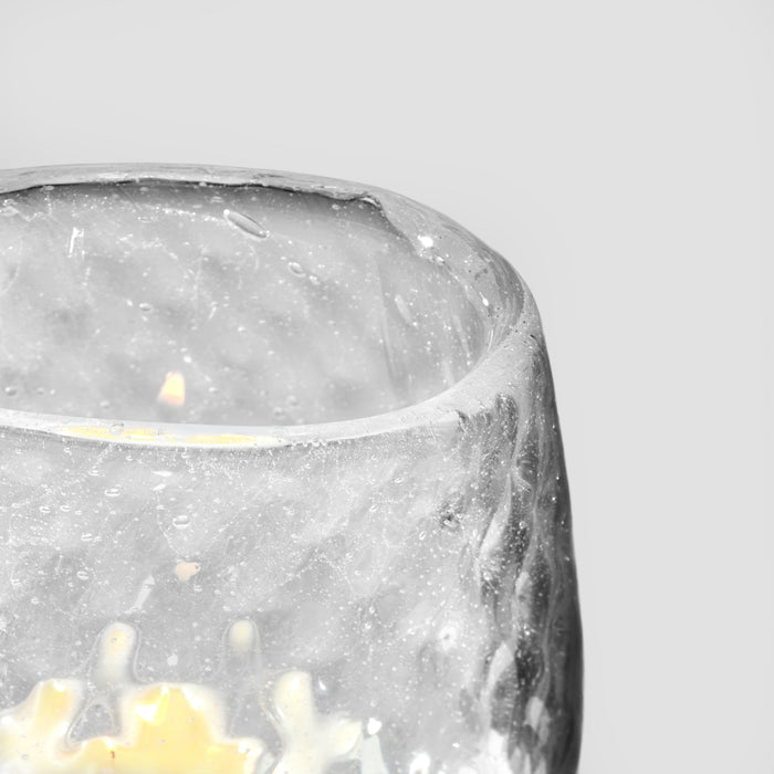 Clear glass tealight holder with speckled pattern containing lit yellow candle.