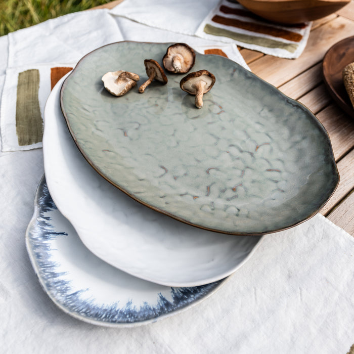 Oval platter with textured gray glaze and brown rim, holding sliced mushrooms, stacked on white plates.