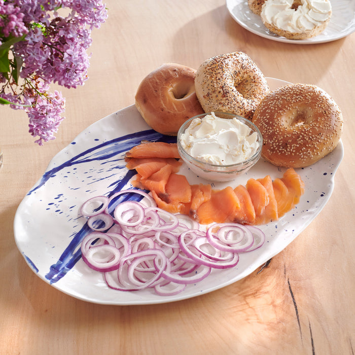 White oval platter with blue brushstroke design holding bagels, cream cheese, smoked salmon, and red onion slices.