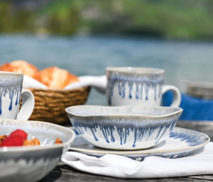 White ceramic tableware with blue brushstroke pattern arranged on table by water with basket of bread.