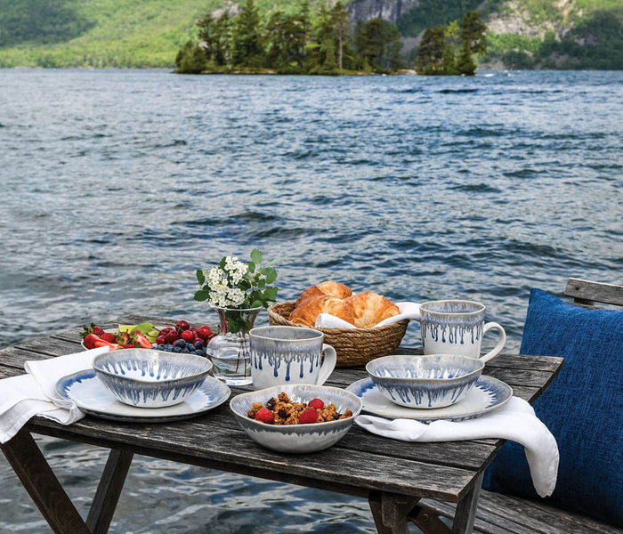 Lakeside table setting with blue-striped dinnerware, fresh berries, croissants, and flowers overlooking forested shoreline.