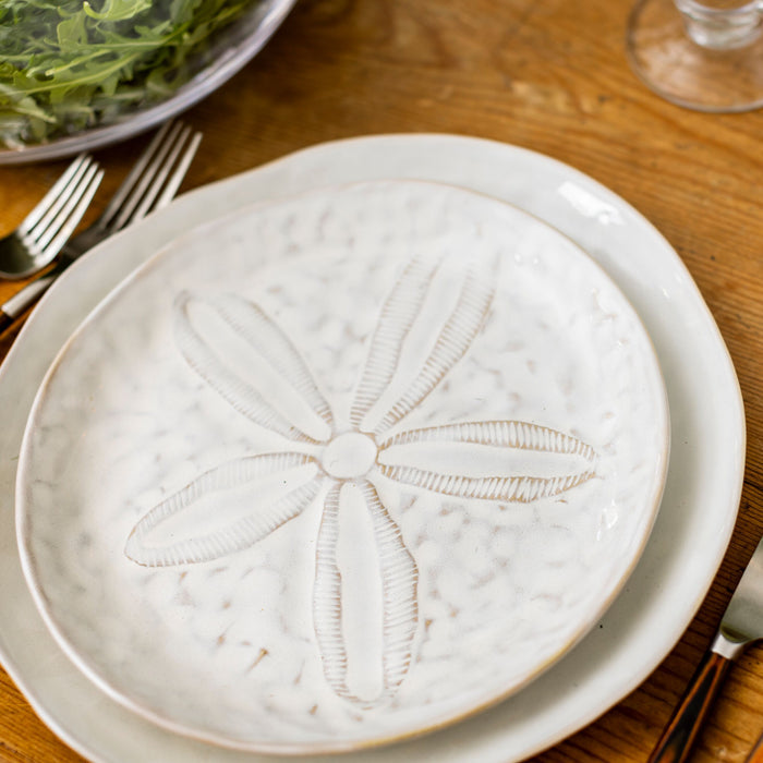 Cream-colored side plate with embossed sand dollar design in center, set on wood table with forks and salad bowl.
