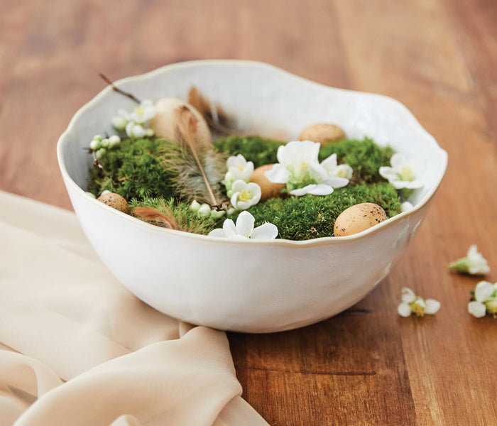 White ceramic bowl with wavy rim containing moss, small white flowers, feathers, and decorative eggs.