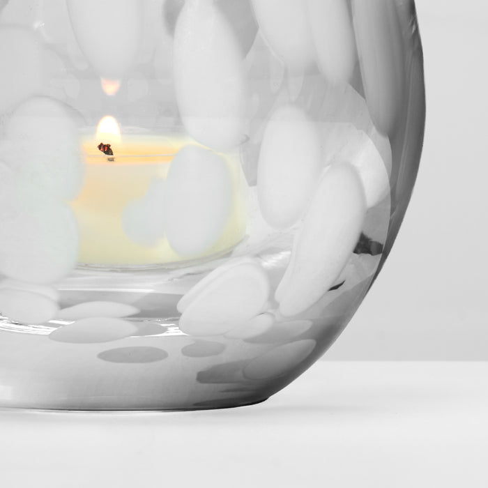 Cream-colored tealight candle with lit flame inside white decorative holder with snowflake pattern.