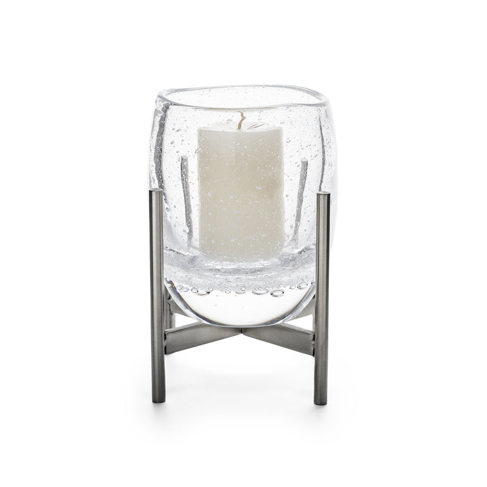 Clear glass hurricane with cream pillar candle inside, supported by stainless steel stand with cross-base design.