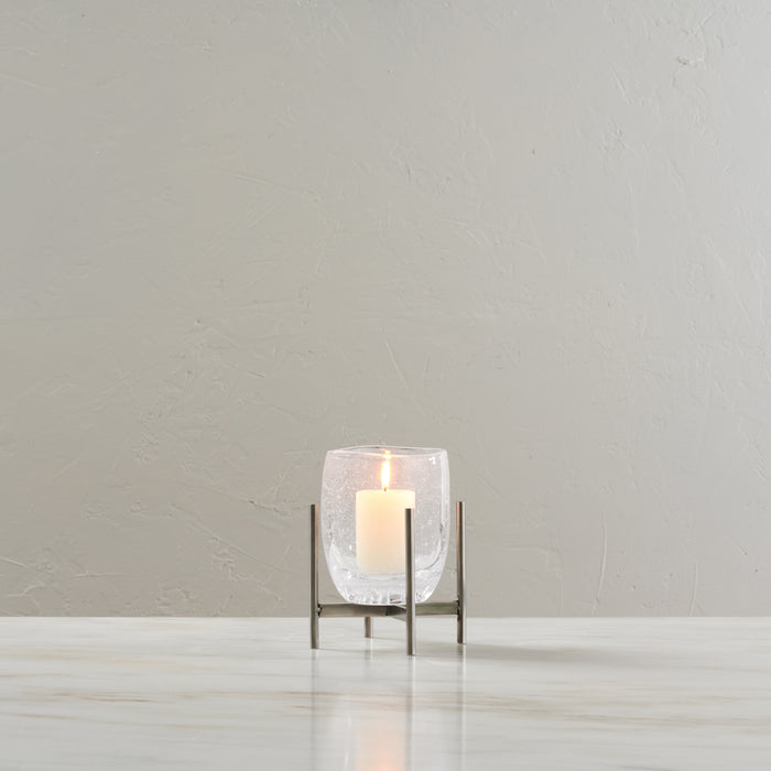 Clear glass hurricane candle holder with stainless steel stand and lit cream candle inside.