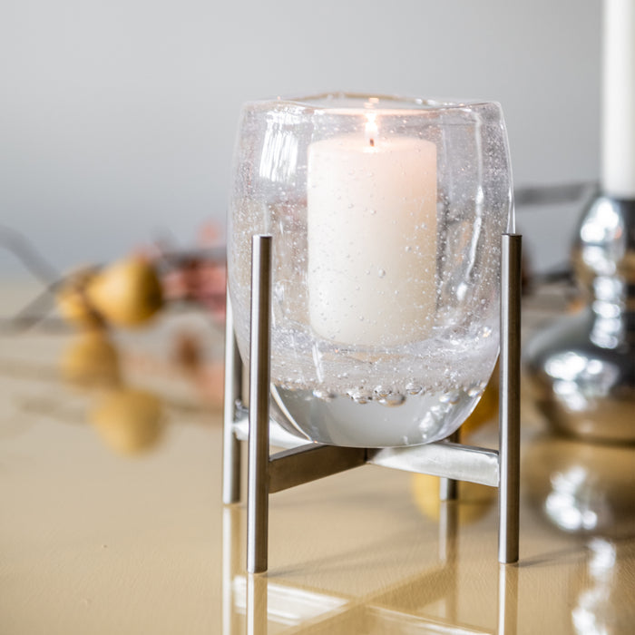 Frosted glass hurricane candle holder with lit cream candle inside stainless steel stand frame.