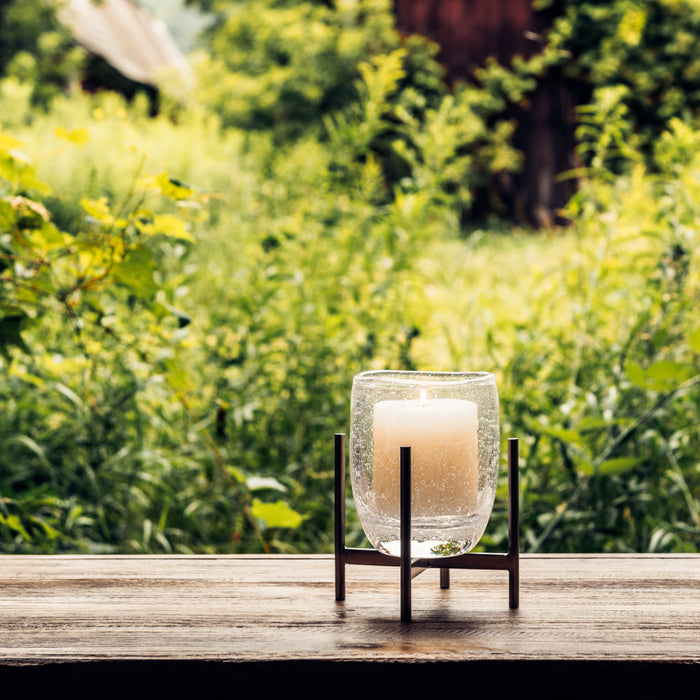 Glass hurricane lantern with cream pillar candle in black stainless steel stand on wooden surface.