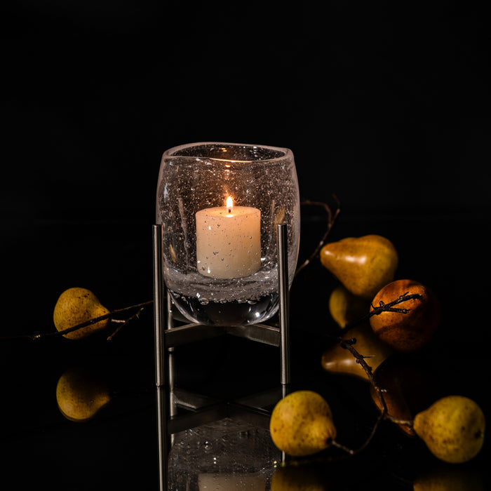 Textured glass hurricane with lit cream candle inside, mounted on stainless steel stand, surrounded by decorative pears and b