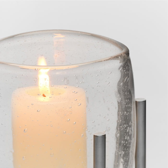 Lit cream candle inside clear glass hurricane with speckled texture and stainless steel stand supports.