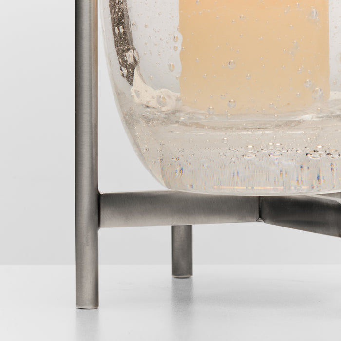 Clear glass hurricane with pale yellow candle on brushed stainless steel stand with support bracket.