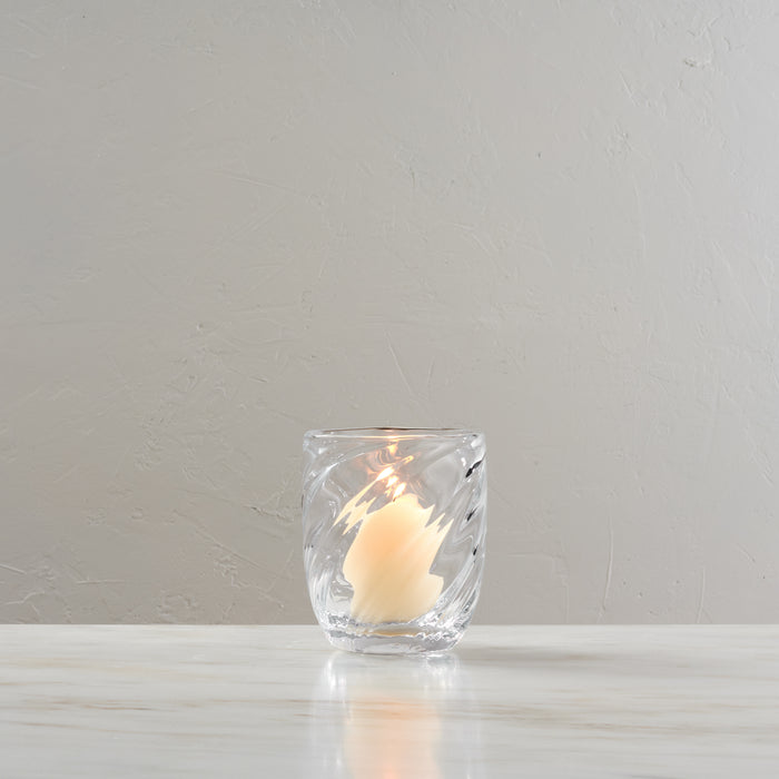 Clear glass votive holder with lit cream-colored candle inside, displayed on white surface.