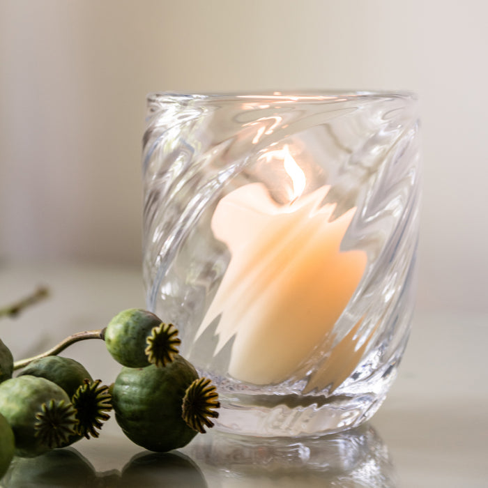 Clear glass candle with cream-colored wax and textured relief pattern, accompanied by green poppy seed pods.