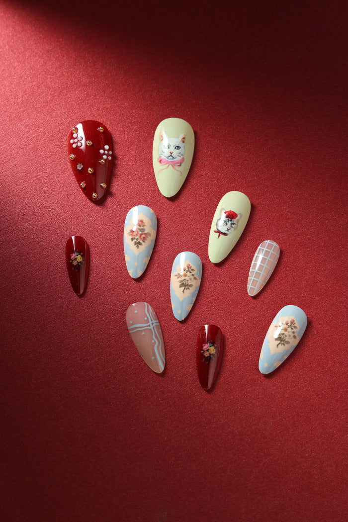 Almond press-on nails featuring a collage of cat designs, floral patterns, and plaid accents in pastel and burgundy colors.