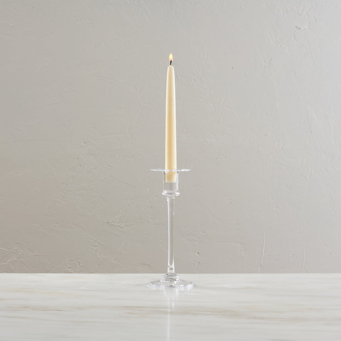 Clear glass candlestick with tall stem holding a lit cream taper candle and horizontal arms.