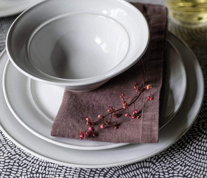 White dinnerware place setting with stacked plates and bowl on grey placemat with brown napkin and berries.